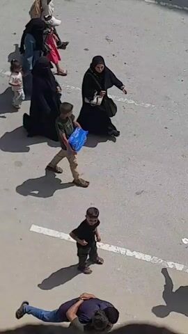 A low-angle aerial view shows a street scene on a sunny day. The pavement is a light gray with a white painted line. In the foreground, two individuals are embracing. Further up the street, a young boy in dark clothing walks with his left hand on his hip. To the left, a group of people, including a woman in a black abaya, are walking away from the camera. To the right, another young boy carries a large blue plastic container. Several other figures, including women in abayas and children, are scattered throughout the scene. Some of the figures are carrying bags or backpacks. Shadows are cast on the pavement, indicating the direction of the sun.