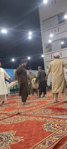Here's a detailed description of the video:

The video shows a group of men performing a traditional dance on a red patterned rug in an outdoor setting. The men are dressed in traditional clothing, mainly long, loose-fitting robes in colors like brown and beige. Some wear caps or head coverings. 

At the center of the group, one man plays a drum, providing the rhythm for the dance. The other men move in a circle, with some holding what appears to be white cloths or handkerchiefs. Their movements involve stepping, turning, and swaying to the beat of the drum.

The setting appears to be a covered outdoor space, possibly a courtyard or open hall. The lighting is provided by a series of bare light bulbs strung overhead. In the background, there's a wall with a painted mural, as well as a glimpse of children sitting and watching the performance. The overall atmosphere suggests a cultural or celebratory event.