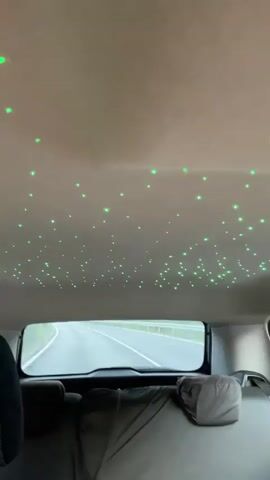 The video displays the interior of a vehicle from the perspective of a rear passenger, looking forward. The car's ceiling is covered with numerous small, illuminated points that cycle through different colors: purple, pink, and green. Through the rear window, a paved road with a yellow dividing line is visible, flanked by green foliage, indicating the car is in motion. A dark-haired person's head and shoulder are partially visible on the left side of the frame. Two grey car headrests and part of a grey car seat are in the foreground.