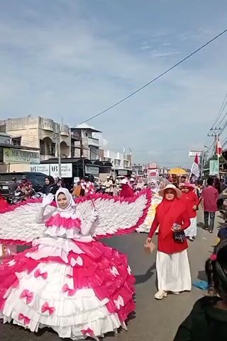 The video shows an outdoor parade on a street lined with buildings. In the foreground, a person wearing a pink and white ruffled dress and a white hijab, with large white wings, waves. Behind them, a woman in a red hijab and a wide-brimmed hat walks forward, holding a small object. Other people, some in red and white clothing and some wearing hijabs, are walking and riding motorcycles in the background. The sky is blue with some clouds.