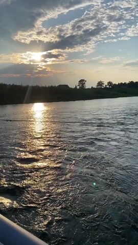 The video captures a serene river scene at sunset in Novosnezhnaya, Russia. The sun, partially obscured by dramatic clouds, casts a golden, shimmering reflection across the water's surface. The river flows gently, its ripples catching the fading light. The left bank of the river is lined with trees and appears to recede into the distance, while the right bank is densely forested with lush green foliage. A white railing is visible in the foreground, suggesting the vantage point might be from a bridge or a dock. The overall mood is peaceful and tranquil, highlighting the beauty of nature at dusk. The time of day is clearly indicated by the low position of the sun and the warm, golden light. The weather appears pleasant, with a mix of sun and clouds creating a dynamic sky.