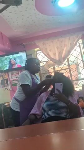 A woman with braided hair and wearing a white T-shirt and purple apron is standing and styling the hair of another woman, who is seated in a chair. The seated woman has dark, afro-textured hair and is wearing a striped shirt and a light pink robe. She is looking at her phone, which she holds up to her face. The scene appears to be inside a small salon, with pink walls and a decorative ceiling. There is a TV on the wall showing a program, and a window with sheer curtains. A large mirror reflects the scene. A brown table or counter is visible in the foreground.