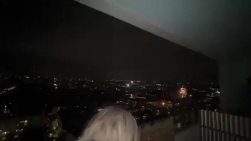 From a high vantage point, possibly a balcony, a nighttime cityscape sprawls below, punctuated by numerous bright lights indicating buildings and streets. Fireworks are sporadically bursting in the distance, creating colorful displays against the dark sky. Initially, two figures, possibly a couple, are seen in the foreground. They are close together, and the video then pans away from them to focus on the fireworks over the city.