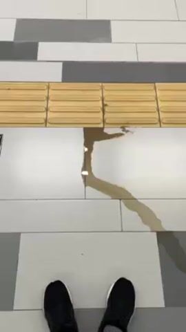 The video shows a person standing on a tiled floor in what appears to be a public space, likely a train station or similar transit hub. The floor is made up of rectangular tiles in alternating shades of white, gray, and a tan color. A row of tactile paving, designed to assist the visually impaired, runs horizontally across the frame. On the floor, there's a noticeable puddle or spill of a dark liquid, potentially coffee or similar drink. It has spread out in a branch-like pattern across the white tiles. The person filming is standing just in front of the spill, wearing black athletic shoes. There are no other people visible in the frame. The video is taken from a first-person perspective, pointing down at the floor.