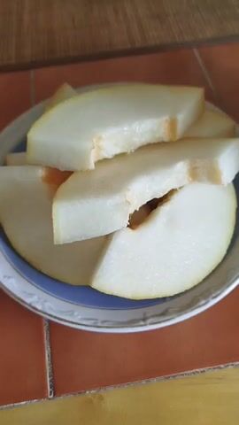 The video shows a close-up of sliced melon on a plate. The plate has a blue and white rim. There are several wedges of pale yellow melon on the plate, with some showing seeds. To the right of the plate is half of a melon, also showing seeds in the center. The melon is sitting on an orange tiled surface. The background is blurred but appears to be a wooden surface with some objects on it. The lighting is natural and appears to be coming from the side.