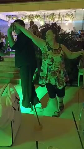 The video shows an outdoor scene at night, likely a party or event. In the foreground, a white bag is on a table, and drinks with straws are visible. The background features a group of people dancing. A woman in a floral dress and black leggings is dancing in the center, with her arms extended. A man in a dark shirt and pants is dancing beside her, also with his arms raised. Other people are dancing in the background. Colored lights, likely from stage lighting, illuminate the area with purple and green hues. Plants and greenery are visible in the background, suggesting an outdoor setting. The overall ambiance appears festive and energetic.