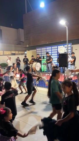 The video shows a live performance of a band, in front of a group of children who are dancing. The band is on a raised stage, with a backdrop promoting “Mall del Sur”.  The band consists of several musicians, including a vocalist, a guitar player, a drummer, a tuba player, a saxophone player, and a percussionist.  The children, both boys and girls of varying ages, are dressed in casual clothing and several carry items that make it appear they are performing a traditional dance.  The performance is taking place in an outdoor area, with palm trees visible in the background and a building in the back with people standing.  The lighting is provided by stage lights and ambient lighting from the surrounding area.