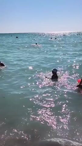 The video shows a group of people swimming in the ocean on a sunny day. The water is clear and blue, with sunlight shimmering on the surface. Several children are playing in the water, some wearing arm floaties. In the foreground, two children are visible, one wearing a blue hat and arm floaties, and another wearing a pink hat and arm floaties. Further out in the ocean, other people can be seen swimming and enjoying the water. The sky is clear and blue, with a few wispy clouds. A bird is seen flying in the distance. The overall impression is one of a peaceful and enjoyable day at the beach.
