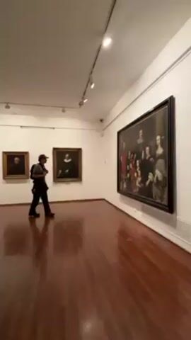 The video shows a man in an art museum. The man, who has a mustache and is wearing a camouflage baseball cap, a gray t-shirt, dark pants, and a black backpack, walks towards the camera in a room with glossy wood floors and white walls. There are three framed paintings on the walls. The painting on the right features a large group portrait. The other two paintings appear to be portraits of individuals. The room has overhead lighting and a security camera mounted on the ceiling. The man stops and looks at the painting to his left.