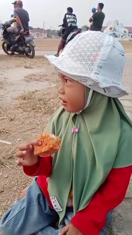 A child wearing a white bucket hat and a green hijab is sitting on the ground and eating a piece of fried chicken. The child is wearing a red long-sleeved shirt and blue jeans. In the background, there are several people and motorcycles. The environment appears to be outdoors, possibly a park or a roadside, with dry grass and dirt on the ground.