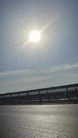 The video shows a view from a moving vehicle, likely a car, driving on a highway. The sun shines brightly in the clear, blue sky with a few wispy clouds. A roadside barrier is visible along the side of the road. Beyond the barrier, there is a line of trees and bushes, passing by as the vehicle moves forward. The pavement is visible in the foreground.