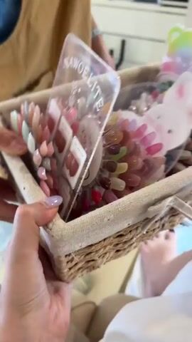 The video displays a close-up of a woven basket filled with an assortment of nail polish samples. The basket is held by a person whose hand, with light purple nail polish, is visible in the foreground. Inside the basket, the nail polish samples are arranged in a circular display, with a clear plastic organizer featuring the brand name "NAWOELITE" at the front. Behind the nail polish, a white decorative cutout of a rabbit with pink ears and cheeks adds a whimsical touch, and a rainbow-colored bear cutout is visible at the top. The nail polish colors range from pastels like pink, lavender, and mint green to warmer tones like peach and coral, as well as some darker shades. Some of the samples are presented in small, square containers labeled "GEL POLISH." The overall scene suggests a nail salon or a personal collection of nail products, likely presented as part of a gift or for selection. The lighting appears to be bright and natural, hinting at a daytime setting, possibly indoors in a well-lit room. The focus is on the colorful variety of nail polish, creating a visually appealing presentation. There are no clear indications of the specific location within Suthep, Thailand, beyond the general context provided. The tone is cheerful and celebratory, evoked by the colorful products and the playful decorative elements.