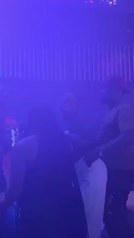 The video shows a group of people dancing in a dimly lit club. The lighting changes colors from pink to green to orange and back to pink. In the foreground, a woman in a black dress with her back to the camera dances. To her right, a woman in a black top and white pants is dancing. To her right, a man wearing a red cap and a brown shirt is also dancing and appears to be interacting with the woman in white pants. In the background, other people are visible, also dancing. There is a table in the center of the frame with bottles and cups on it. The walls of the club are lined with vertical striped panels that are illuminated by the changing lights.