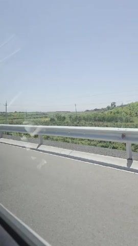 The video shows a bright, sunny daytime view from inside a moving vehicle, possibly a car or bus, in Kosh, Armenia. The scene captures the landscape alongside a paved road, looking over a metal guardrail. Beyond the guardrail, there are open fields with green vegetation, possibly agricultural land or natural foliage, extending towards a distant horizon. Several power lines and at least two power pylons are visible against the clear, light blue sky. The road surface is grey asphalt, and a concrete curb runs along the base of the guardrail. Reflections of the vehicle's interior or windows can be seen on the left side of the frame.