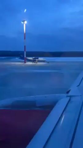 The video shows an airport scene at dusk. From inside a passenger plane, the viewer sees the wing of their plane, painted white and red, through the window. On the tarmac, another smaller turboprop plane sits under a bright light on a pole painted red and white. The sky is a deep blue with some clouds. The ground has patches of snow. There are no people visible in the video.