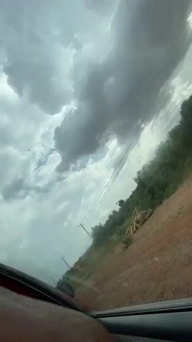 The video depicts a scene viewed from inside a vehicle, likely a car, moving along a dirt road. The perspective is angled upwards, emphasizing the sky dominated by dark, ominous clouds, which suggest an approaching storm. Power lines run parallel to the road, supported by wooden poles. The landscape adjacent to the road features a grassy embankment and some trees. The windshield of the car has some visible smudges. The color palette is muted, reflecting the overcast weather conditions.
