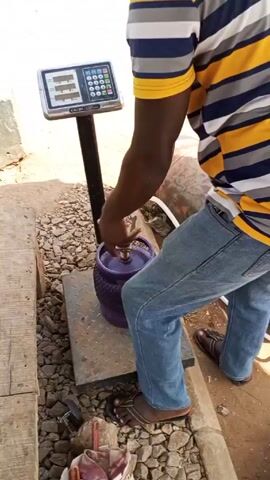 The video shows a man refilling a gas cylinder. He's standing in an outdoor area, likely a gas station or refill point, with a purple gas cylinder on a digital weighing scale. A white hose connects the cylinder to a larger, horizontal tank in the background. The man is wearing a striped shirt and jeans. He adjusts the hose connection before taking a step back. The scale has a keypad and a digital display. The area around the scale appears to be a mix of concrete and gravel.