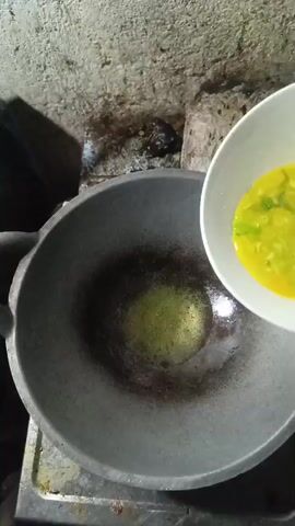 A bowl of beaten eggs with green onions is being poured into a hot wok with oil. The wok is black and appears to be cast iron. The background is a rough, textured wall and some objects that are out of focus. The lighting is dim.