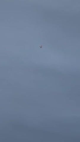 A small airplane is visible in the center of the frame, flying against a pale blue sky with scattered clouds. The airplane appears to be a propeller-driven aircraft, with its wings and tail clearly distinguishable. The camera is positioned below the airplane, looking upwards. The airplane is stationary in the frame for the duration of the video. The lighting suggests it is daytime, possibly overcast or early morning/late afternoon.