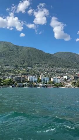The video begins with a view of a body of water with moderate waves. A coastline with buildings and a densely populated hillside covered in trees and houses is visible in the background. A range of mountains forms the backdrop to the scene. The sky is blue with scattered white clouds. The camera appears to be on a moving boat, indicated by the water's motion and the presence of a metal railing in the foreground. The video is shot in landscape orientation. The lighting suggests it is daytime.