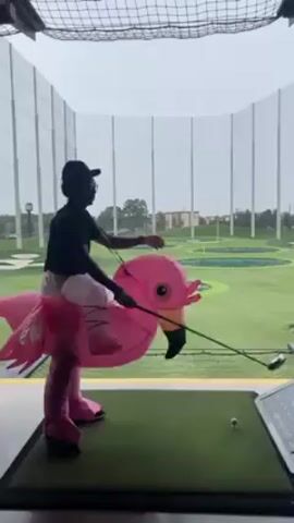 A person wearing an inflatable pink flamingo costume is at an indoor golf driving range, swinging a golf club at a golf ball. The person is wearing a black shirt, light-colored pants, and a black cap. They are positioned on a green artificial turf mat with a golf ball teed up. The driving range is enclosed by a net, and the outdoor area visible through the net includes a green golf course. The weather appears overcast or rainy.