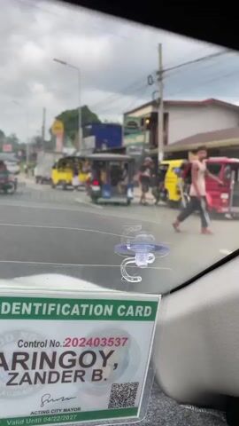 The video shows a street in what appears to be a developing area. The view is from inside a vehicle, through the windshield, with a driver's identification card visible in the lower left corner. The ID card has a photo of the driver and the name "ARINGOY, ZANDER B.". Outside, the street features a mix of vehicles including motorcycles, tuk-tuks (small motorized rickshaws), cars, and trucks. Some people are walking along the roadside or standing near vehicles. The buildings lining the street are small, and some have commercial signs. The sky is overcast, and utility poles with power lines are visible overhead. The video seems to have been shot during the daytime, with moderate traffic on the road.