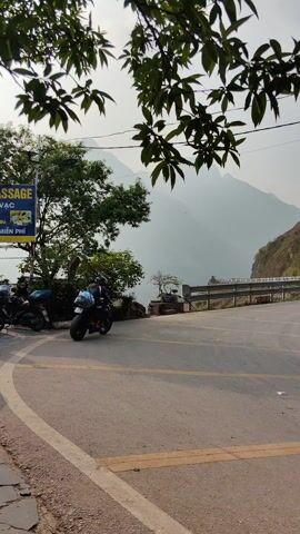 A motorcycle speeds around a bend in a winding mountain road. The road is lined with yellow stripes. In the background, hazy mountains rise into the sky. A sign reading "MASSAGE" in Vietnamese is visible on the left. As the motorcycle moves out of view, a man in a white shirt and dark shorts walks across the yellow stripes, heading towards a parked silver sedan and a white van. A few other vehicles are parked further up the road. Overhead power lines are visible. The scene is framed by tree branches in the foreground.