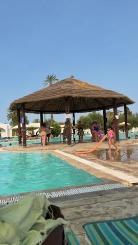 The video opens on a bright, sunny day with a clear blue sky. In the foreground, a pool with turquoise water is visible, with a striped lounge chair in the lower right corner. The main focus of the video is a thatched-roof gazebo situated over a paved area beside the pool. Several people are gathered under and around the gazebo. A young girl in a pink swimsuit is holding a long, orange inflatable tube and appears to be interacting with it near the edge of the pool. Other individuals, dressed in swimwear and casual clothing, are standing and talking within the shaded area of the gazebo. Palm trees and some vegetation are visible in the background, suggesting a resort or outdoor recreational setting. The overall atmosphere appears relaxed and casual, consistent with a day at a swimming pool.