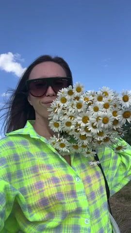 A woman wearing sunglasses and a neon green and purple plaid shirt stands in a field, holding a large bouquet of white daisies in front of her face. The sky is blue with some white clouds, and trees are visible in the background. She appears to be moving and looking at the camera.