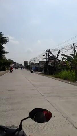 A first-person perspective video shows a person riding a motorcycle down a paved road on a sunny day. The road is lined with buildings, trees, and utility poles with many wires. Several other motorcycles are visible on the road, some passing the rider, others being passed. The rider's hand and a portion of the motorcycle's mirror are visible in the foreground. The sky is mostly clear with some scattered clouds.