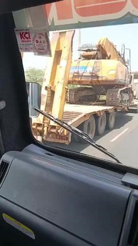 The video shows a view from the inside of a truck driving on a highway. The truck is following a flatbed truck carrying a large yellow excavator. The flatbed truck has multiple axles.  The excavator is secured to the flatbed with straps. The sky is bright and sunny. There are other trucks on the road in the distance.  The dashboard and windshield wipers of the truck are visible.  A warranty sticker is visible in the top left corner of the windshield.