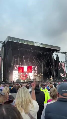 A wide shot captures an outdoor concert stage with a large screen displaying a person playing a guitar. The stage is illuminated by bright lights, and the "SUMMER SESSIONS" logo is visible. A large crowd of people, seen from behind, stands and sits in front of the stage, looking towards the performance. The sky is overcast.