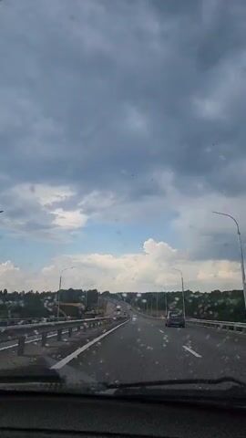A view from inside a vehicle driving on a highway. The road is wet with what appears to be raindrops on the windshield. The sky is cloudy with patches of blue sky visible. There are trees and construction equipment on the side of the road. Several other vehicles are on the highway, including cars and trucks. The video is shot from a first-person perspective, looking forward through the windshield.