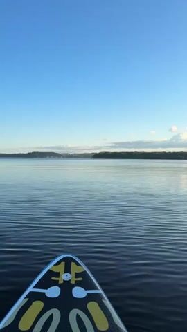 The video shows a view from a paddleboard on a calm lake under a clear blue sky. The front of the paddleboard, decorated with a yellow and black pattern, is visible in the foreground. The water has gentle ripples, reflecting the sky. In the distance, a tree-lined shore is visible, with a few scattered clouds in the sky. The scene depicts a serene outdoor environment.