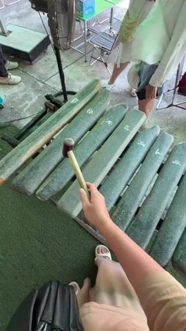 The video shows a first-person perspective of someone playing a large, stone xylophone. The xylophone has many grey, rectangular stone bars of varying lengths. The player is holding a mallet with a wooden handle and a brown rubber head and striking the bars. The sound of the xylophone can be heard. In the background, several people are visible. To the left, a person wearing a white shirt and dark pants is standing near a fan. In the center background, a person in a red shirt is seated at a table with papers. To the right, another person, partially visible, is also seated at a table. The environment appears to be outdoors, possibly in a recreational area or an event, with a green artificial turf on the ground and a concrete or paved area around the xylophone. The lighting suggests it is daytime.