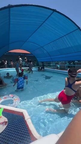 This video captures a lively scene at an outdoor swimming pool in Kiryat Yam, Israel. The pool is covered by a large, blue canopy, suggesting a sunny day with a need for shade. Several people, primarily children and some adults, are enjoying the water.

In the foreground, a young girl in a red swimsuit with a blue top is partially submerged, her legs extended out of the water. She appears to be playing or posing for the camera. To her left, a young boy in a blue swimsuit with a character print is standing in the shallow end, holding onto an inflatable ring. Other children are visible in the pool, some with inflatable toys, splashing and playing. In the background, a group of people, including women and children, are gathered near the edge of the pool, some appearing to be supervising.

The overall atmosphere seems joyful and relaxed, indicative of a summer day at a public or resort pool. The water is clear and inviting, reflecting the bright sunlight. The setting appears to be a recreational area, likely within Kiryat Yam, given the context. There is no visible text within the video.