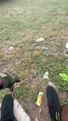 Here's a description of the video:

The video shows a grassy field, with a group of people in the background. Some of them appear to be standing while others are sitting or lying down.  There is a building or wall in the background, as well as some trees. The video is shot from a low perspective, showing the feet and legs of the person recording. They're wearing dark athletic shoes, green socks, and what looks like athletic pants or shorts. There is trash on the ground near the camera. The lighting suggests it is either early morning or late afternoon. The person recording appears to be sitting still. There is a bit of movement from the camera as the person recording shifts position.