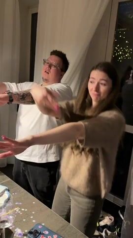 Three people are in a kitchen. Two are dancing to some music with their arms outstretched. The third person appears to be offering a bottle of champagne. The table is covered in glasses, confetti, and a phone. There is a window with string lights in the background.