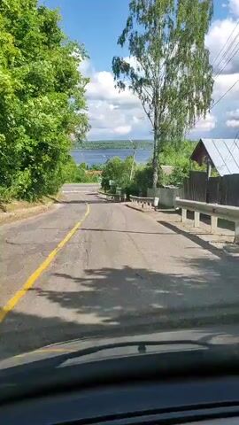 The video shows a point-of-view perspective from inside a vehicle, looking out at a road that slopes downhill. The road is paved and has a yellow line marking the lane. On either side of the road, there are trees and vegetation. In the distance, a body of water, likely a lake or river, is visible under a partly cloudy sky. There are also some houses and fences along the right side of the road. The sun is shining, casting shadows on the road. There are no people visible in the frame.