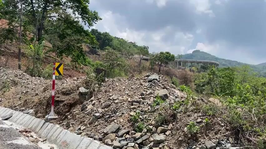 The video shows a hillside with a road at the bottom and a concrete overpass in the distance. The hillside is covered with rocks, dirt, and some vegetation. A road sign is visible next to the road. A banana plant is at the bottom right of the frame. The sky is cloudy.