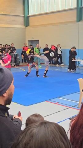 The video shows two men engaging in a kickboxing match on a blue mat in a gymnasium. They are wearing protective headgear and gloves. The man on the left has yellow accents on his headgear, while the one on the right has red. Spectators are visible on the sidelines, observing the match. A table with judges and scorekeeping equipment is set up near one edge of the mat. The environment includes gymnasium markings on the floor and light-colored walls with windows.