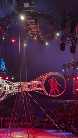 The video shows a circus performance in a large tent. In the center of the frame is a complex apparatus consisting of two large metal wheels connected by a central structure. Two performers, dressed in matching red and white costumes, are inside one of the wheels, facing each other. They appear to be interacting or performing a stunt. Another performer, also in a similar costume, is visible inside the second wheel on the left side of the frame. The wheels are suspended from a circular rig above, which is illuminated by various colored lights, predominantly red and blue. The background shows rows of seats filled with an audience, and the overall atmosphere suggests a live entertainment event. The lighting is dramatic, with spotlights and stage lights creating a theatrical effect. The camera is positioned relatively close to the performers, capturing the intricate details of the apparatus and the performers' actions. The video captures a dynamic moment of the performance, highlighting the acrobatic nature of the act.