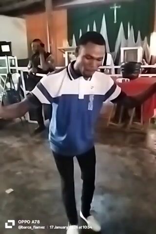A man dances in the center of a room with a concrete floor. He wears a short-sleeved polo shirt that is blue with white stripes, black pants, and white shoes. He holds a black object in his right hand. His arms are extended to the sides. Behind him, another man stands by a microphone stand and sings. Behind them is a table covered with a red cloth, and a backdrop of green and white fabric with a white cross. To the left of the main dancer are some chairs and equipment. The words “OPPO A78 @barca_flamez 17 January 2026 at 10:34” are at the bottom of the screen.
