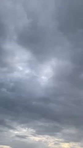 A vertical video shows a dark, overcast sky with scattered patches of lighter clouds. A single thin, dark, vertical object, possibly a piece of wood or a pole, enters the frame from the right and extends upwards. In the lower part of the frame, the indistinct outline of a roof structure is visible against a brighter, yellowish-tinged horizon. The overall impression is of a cloudy, possibly impending weather event.