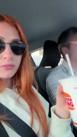 A person with red hair and sunglasses films the interior of a car, where a person with short graying hair and a gray and white plaid shirt drives. Both appear to be looking ahead out of the car, and a cup with a straw and the Dunkin’ logo is shown. The car is moving.
