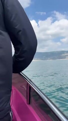 The video shows a boat moving on the water towards land. The boat is pink with a metal railing. A person's hand is holding the railing. The water is a greenish blue, and there are waves. The sky is blue with puffy white clouds. The land in the background has low mountains, some sandy colored patches and some green patches. The day appears sunny and breezy.