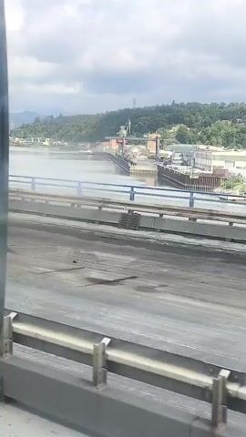The video captures a view from a moving vehicle, likely a train, looking out a window. The foreground shows the interior of the vehicle, with a glimpse of a side panel and the top of a seat. The middle ground features a bridge with concrete barriers and a blue railing. Beyond the bridge, a wide river flows, bordered by a tree-covered hillside. On the opposite bank of the river, a harbor or industrial area is visible, with various buildings, cranes, and what appear to be cargo containers. The sky is cloudy. The overall impression is of a scenic view from a journey, with a mix of natural landscape and industrial activity.