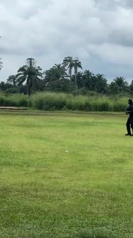 The video shows a person in black walking across a grassy field. The person is wearing what appears to be a uniform and carrying a weapon. In the background, there are palm trees and a metal tower-like structure. A black vehicle is parked on the left side of the frame. The sky is overcast.