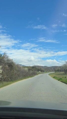 A car is driving down a narrow, cracked asphalt road on a sunny day with a blue sky dotted with fluffy white clouds. Sparse vegetation flanks both sides of the road, giving way to rolling hills in the distance. Power lines are visible along the roadside. The scene is captured from inside the car, with the dashboard visible at the bottom of the frame.