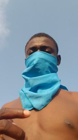 A shirtless, dark-skinned person with short hair looks down toward the camera. Their face is partially covered by a light blue scarf. The background is a clear, light blue sky. The person is likely standing outdoors.